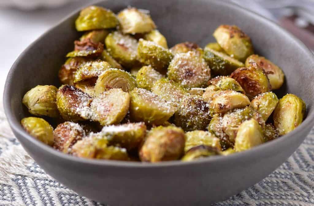 A gray bowl filled with roasted Brussels sprouts sprinkled with grated cheese, placed on a woven placemat.