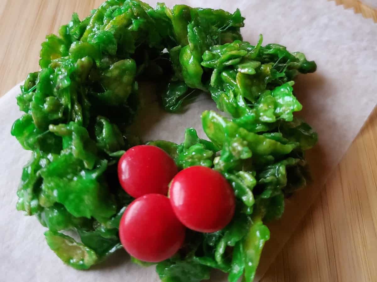 A green cornflake wreath cookie decorated with three red candy pieces, placed on a piece of parchment paper on a wooden surface.