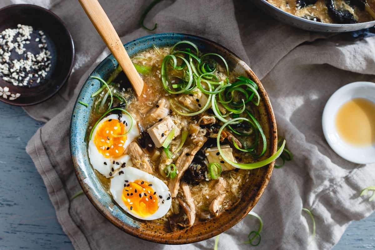 A bowl of ramen topped with a soft-boiled egg, sliced scallions, mushrooms, tofu, and sesame seeds, with broth and a wooden spoon.