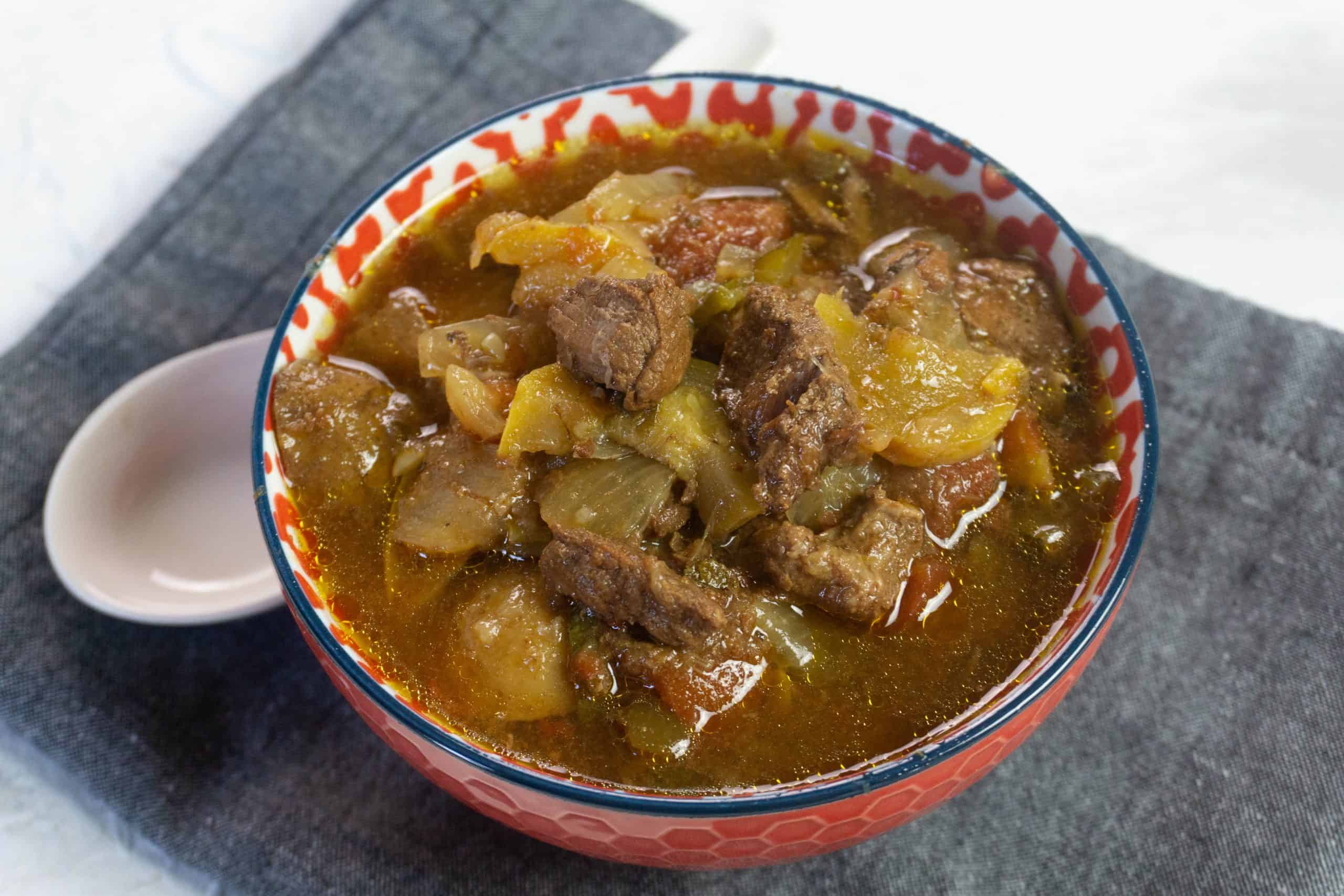 A bowl of beef stew with vegetables in a red-patterned bowl next to a white spoon, placed on a dark cloth napkin.
