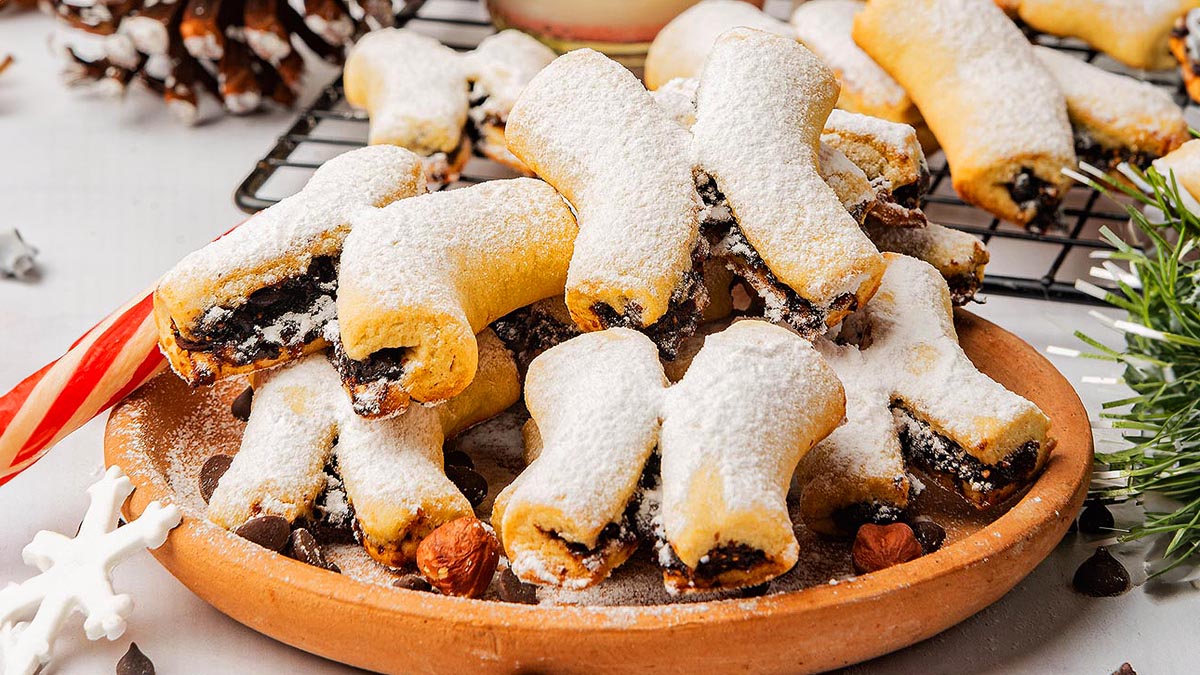 A plate of fig-filled cookies dusted with powdered sugar, with more cookies cooling on a rack in the background. Holiday decorations and candy cane visible nearby.