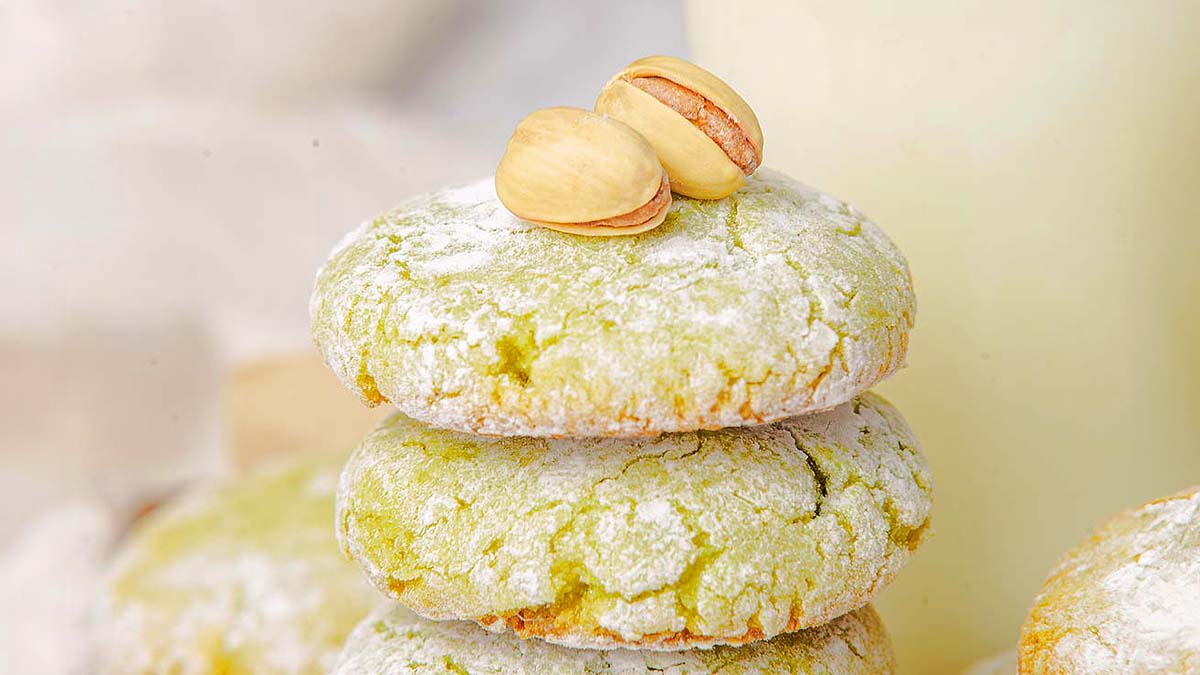 Three pistachio cookies dusted with powdered sugar are stacked, with two pistachio nuts placed on top.