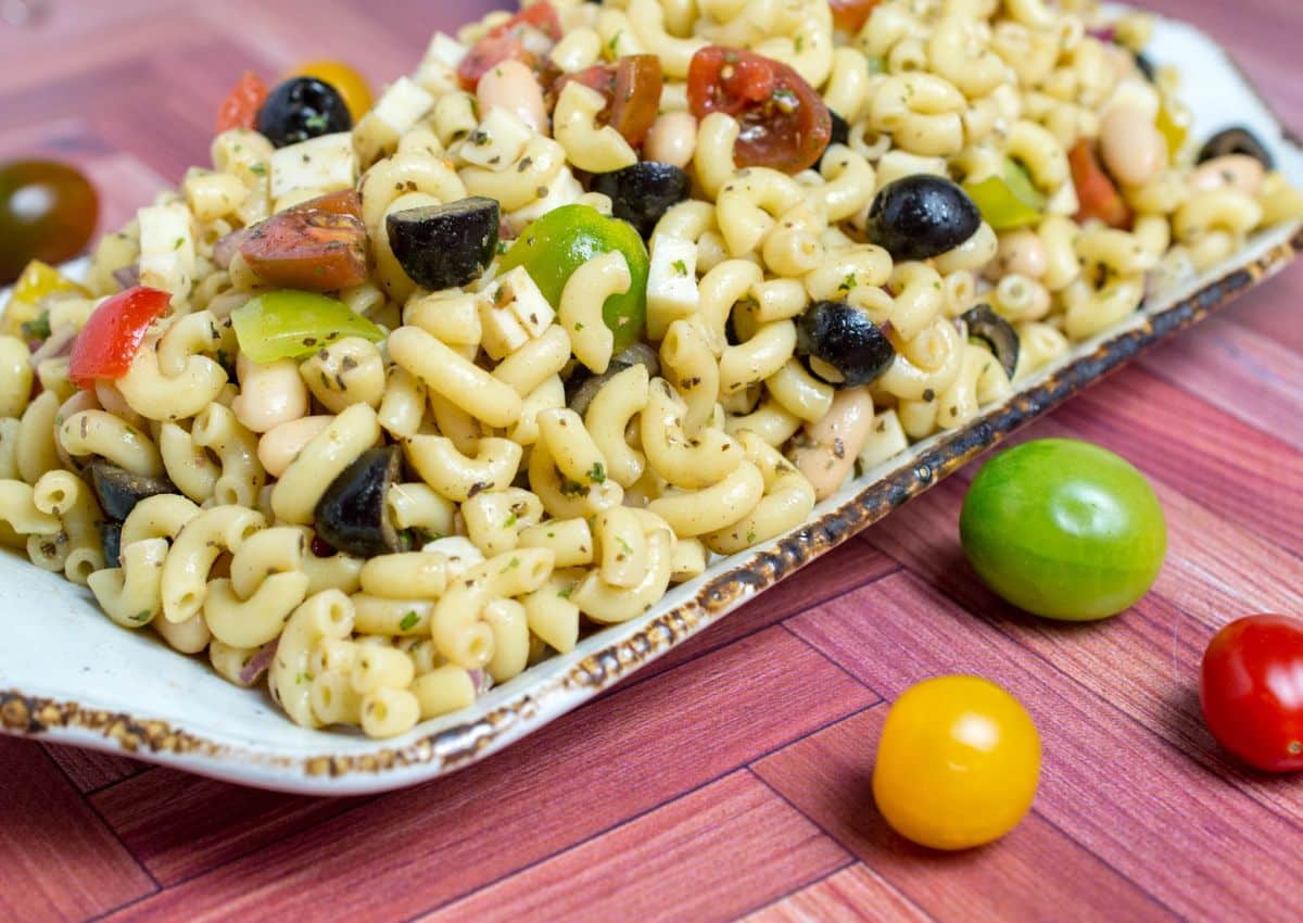 A plate of macaroni pasta salad with black olives, green bell peppers, diced tomatoes, and herbs sits on a wooden surface next to cherry tomatoes.