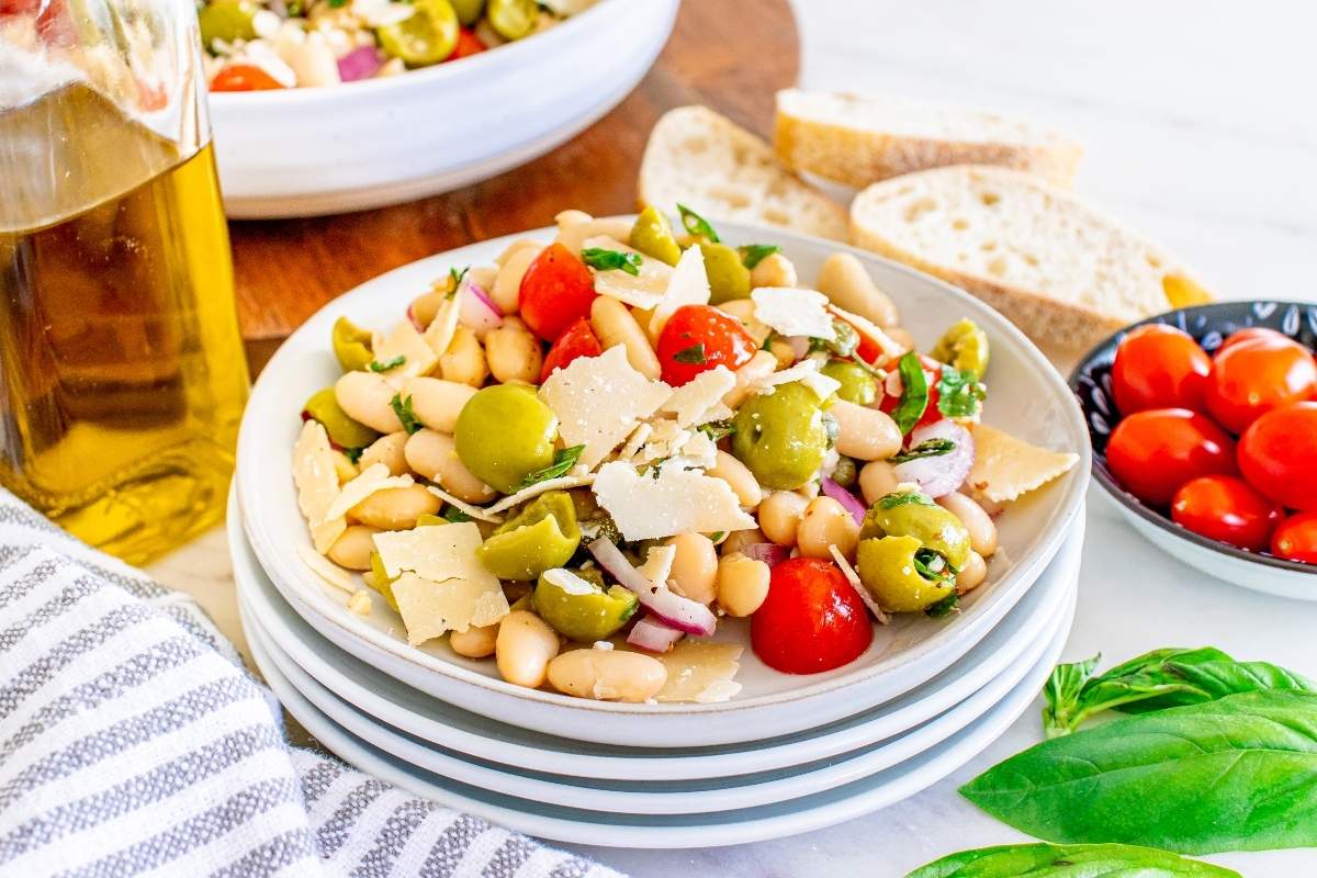 A plate of white bean salad with cherry tomatoes, green olives, red onion, parsley, and shaved cheese, served with slices of bread and a bottle of olive oil nearby.