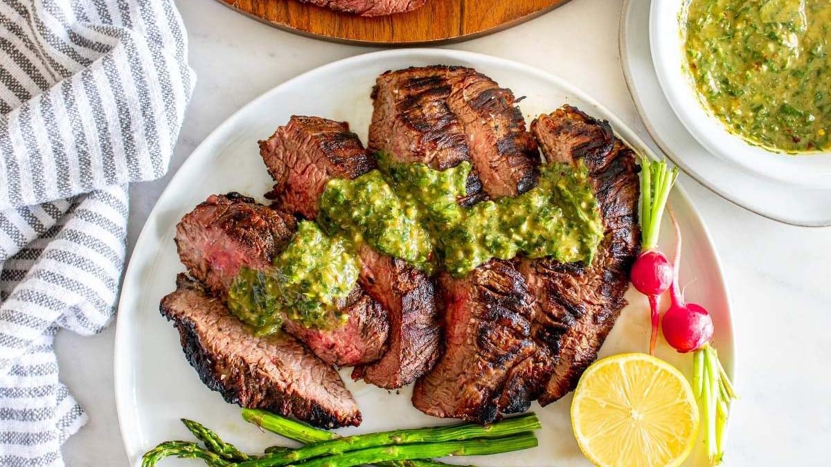 Grilled steak slices topped with green sauce, served with asparagus, radishes, and a lemon half on a white plate.
