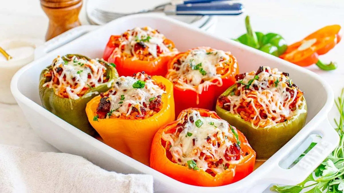 A white baking dish filled with colorful bell peppers stuffed with rice, vegetables, and topped with melted cheese, garnished with chopped herbs.