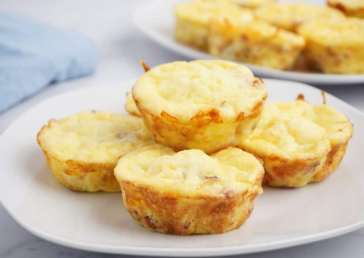 A white plate with several baked egg muffins, golden on the edges, stacked in the center. More egg muffins are visible on a plate in the background.