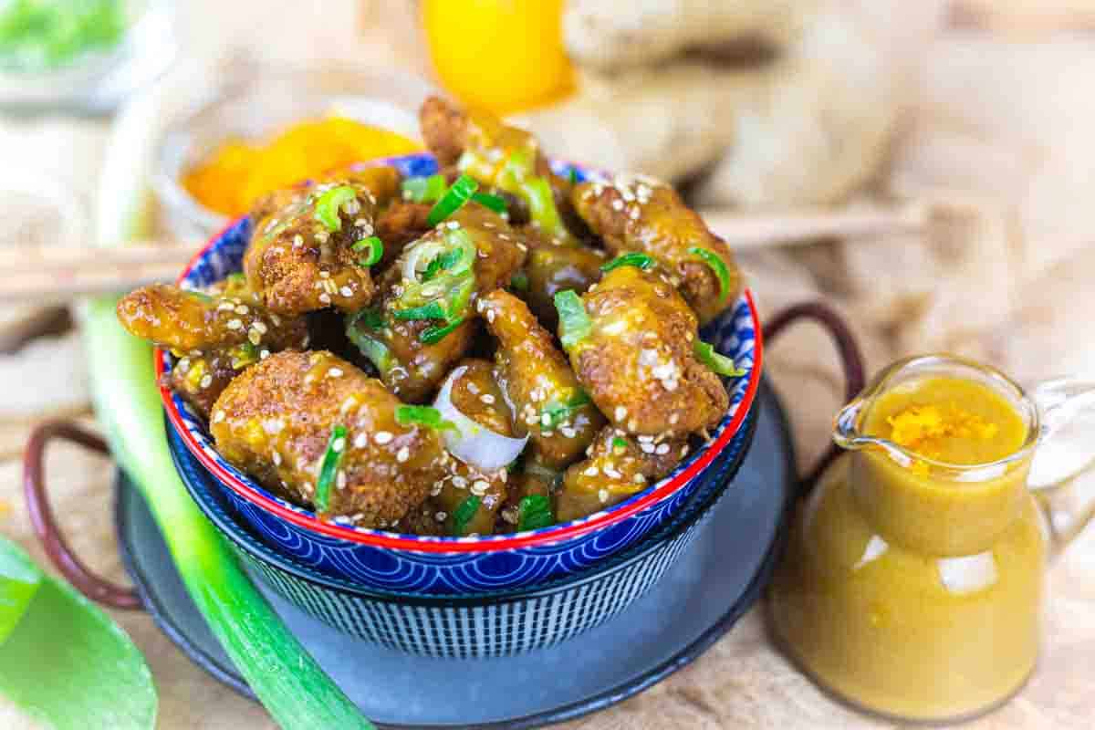 A bowl of crispy golden cauliflower bites garnished with sesame seeds and green onions, served with a side of yellow dipping sauce.