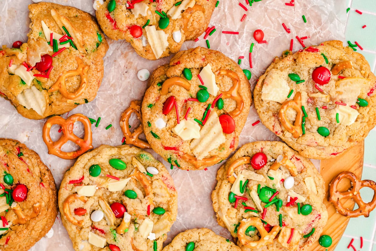 Cookies topped with potato chips, pretzels, red and green chocolate candies, sprinkles, and white chocolate chips, arranged on parchment paper.