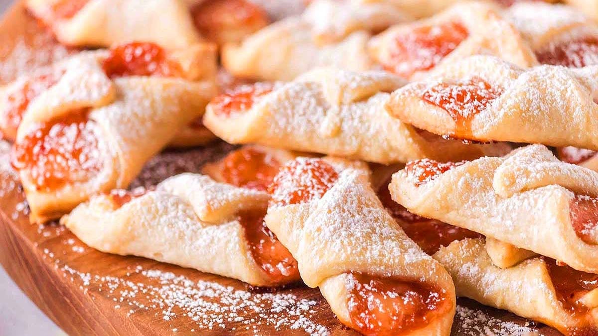 A pile of folded pastry cookies filled with fruit jam and dusted with powdered sugar, arranged on a wooden surface.