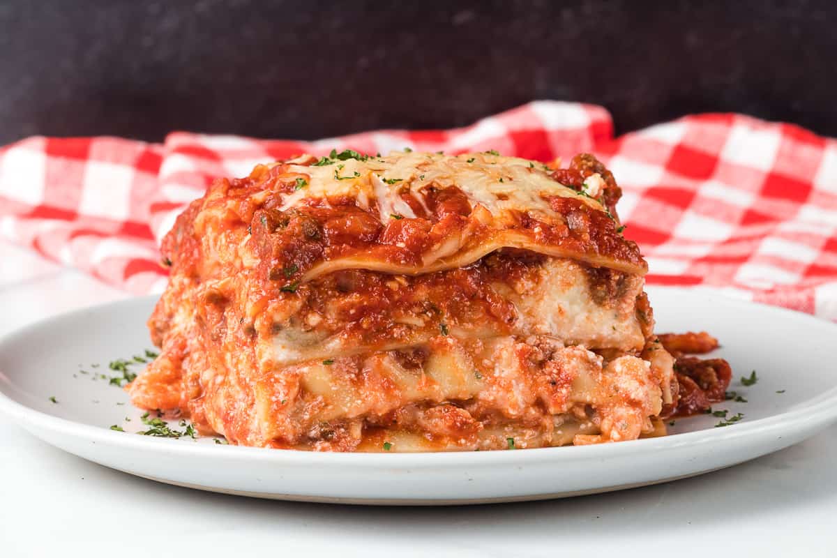 A slice of lasagna with layers of pasta, meat sauce, and cheese, served on a white plate with a red and white checkered cloth in the background.