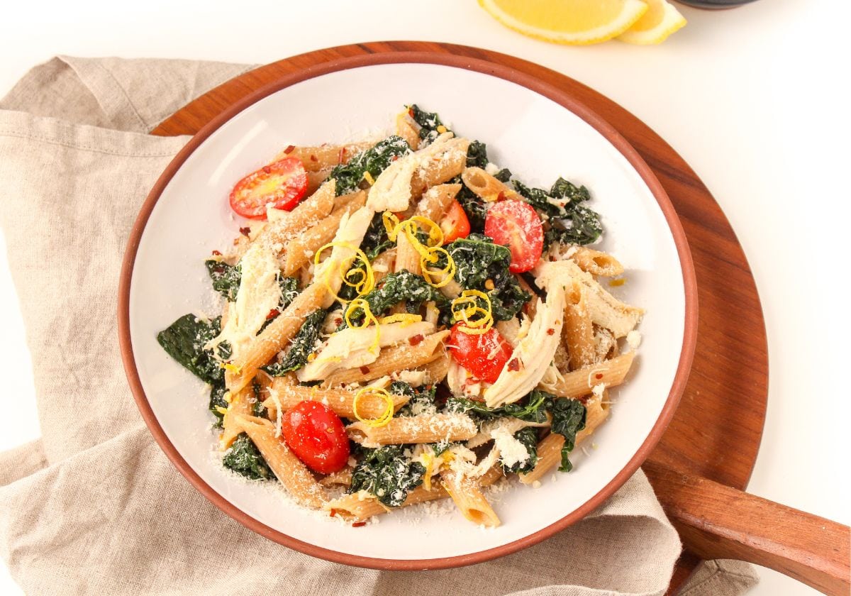 A plate of penne pasta with shredded chicken, kale, cherry tomatoes, grated cheese, and lemon zest, served on a wooden board with a linen napkin and lemon wedges.