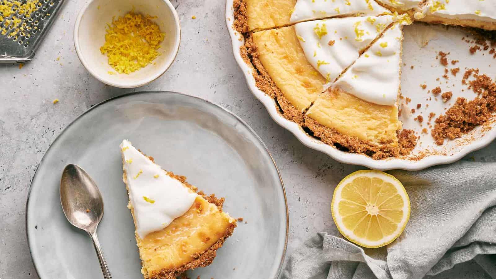 A slice of lemon pie with whipped topping on a plate next to a spoon, with the remaining pie, a halved lemon, and lemon zest in bowls nearby.