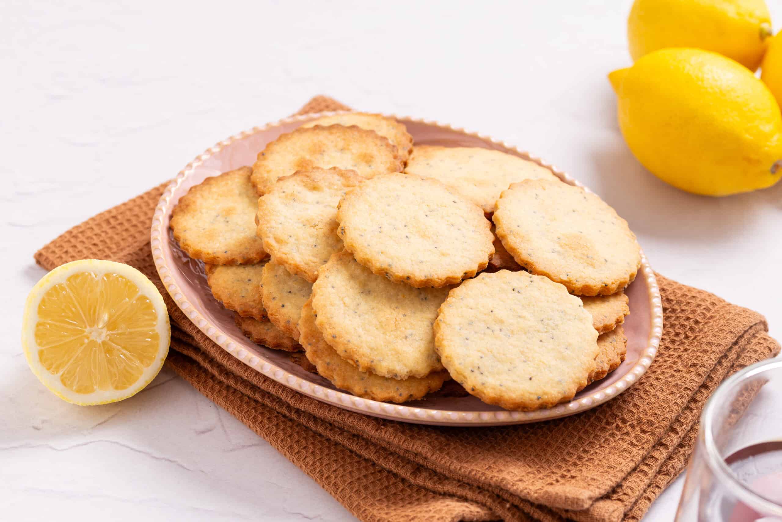 A plate of round, golden cookies sits on a brown towel, with whole and halved lemons nearby on a white surface.