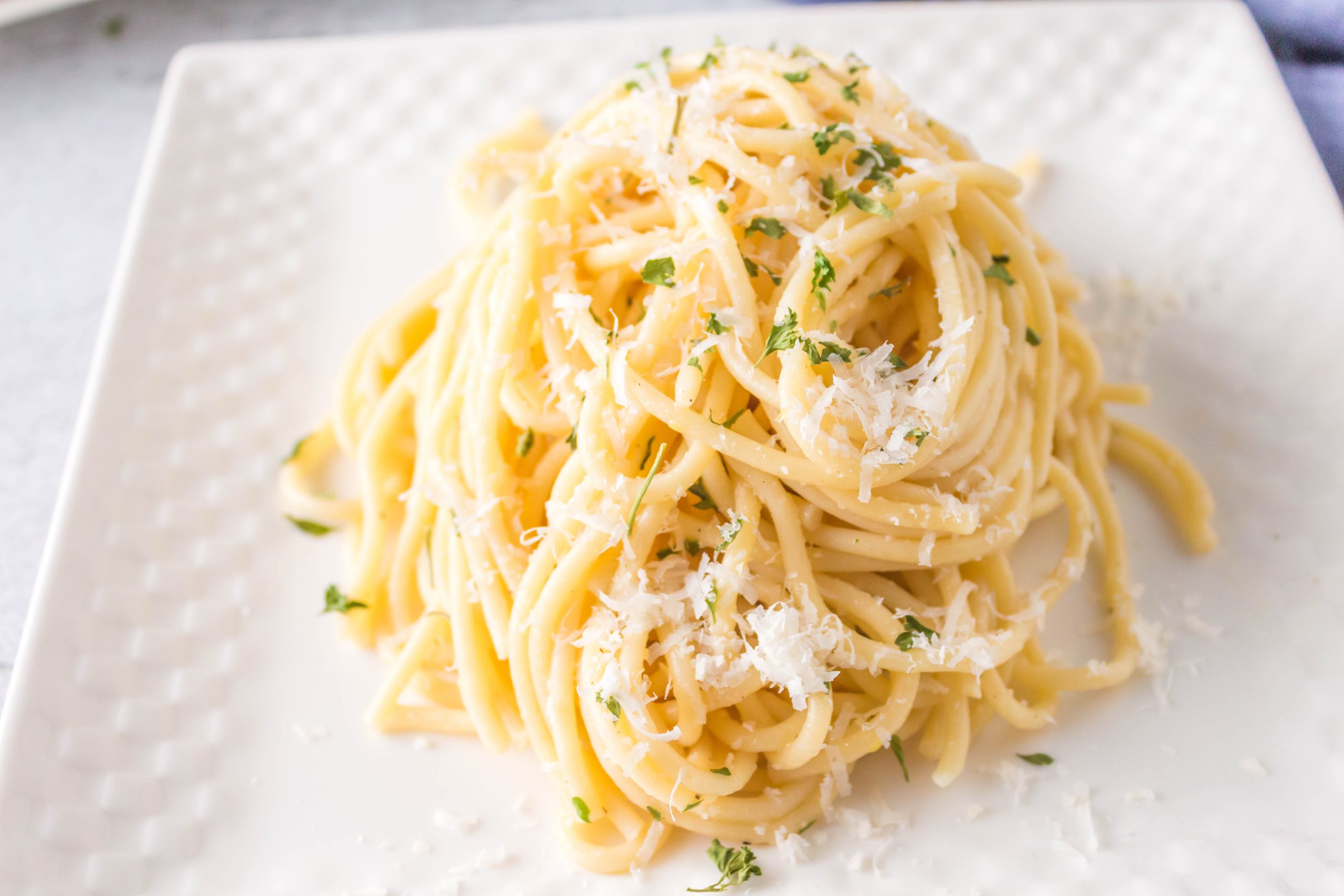 A serving of spaghetti topped with grated cheese and chopped herbs on a white square plate.
