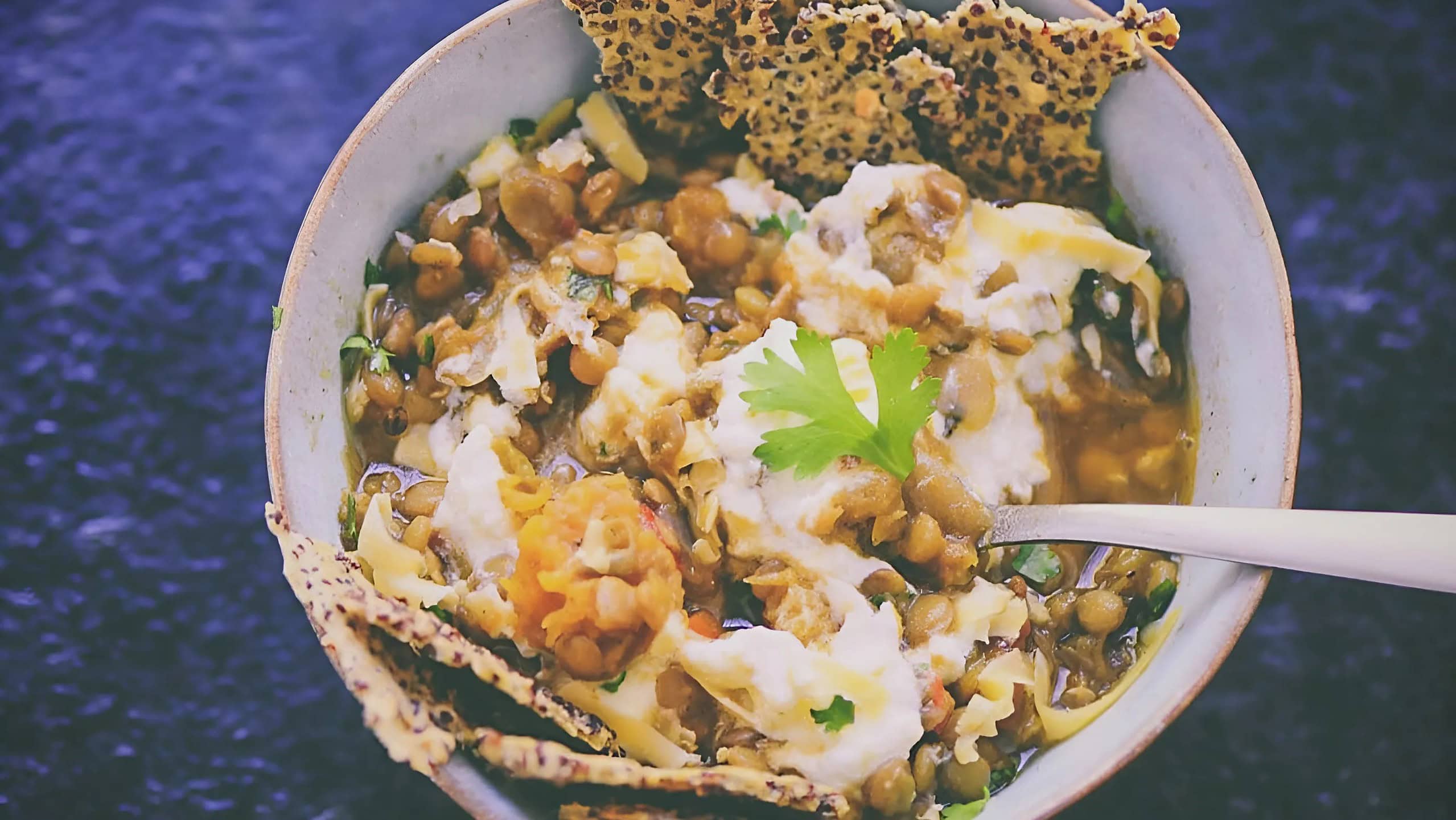 A bowl of lentil stew topped with yogurt and a sprig of cilantro, served with crisp flatbread pieces on the side and a spoon.