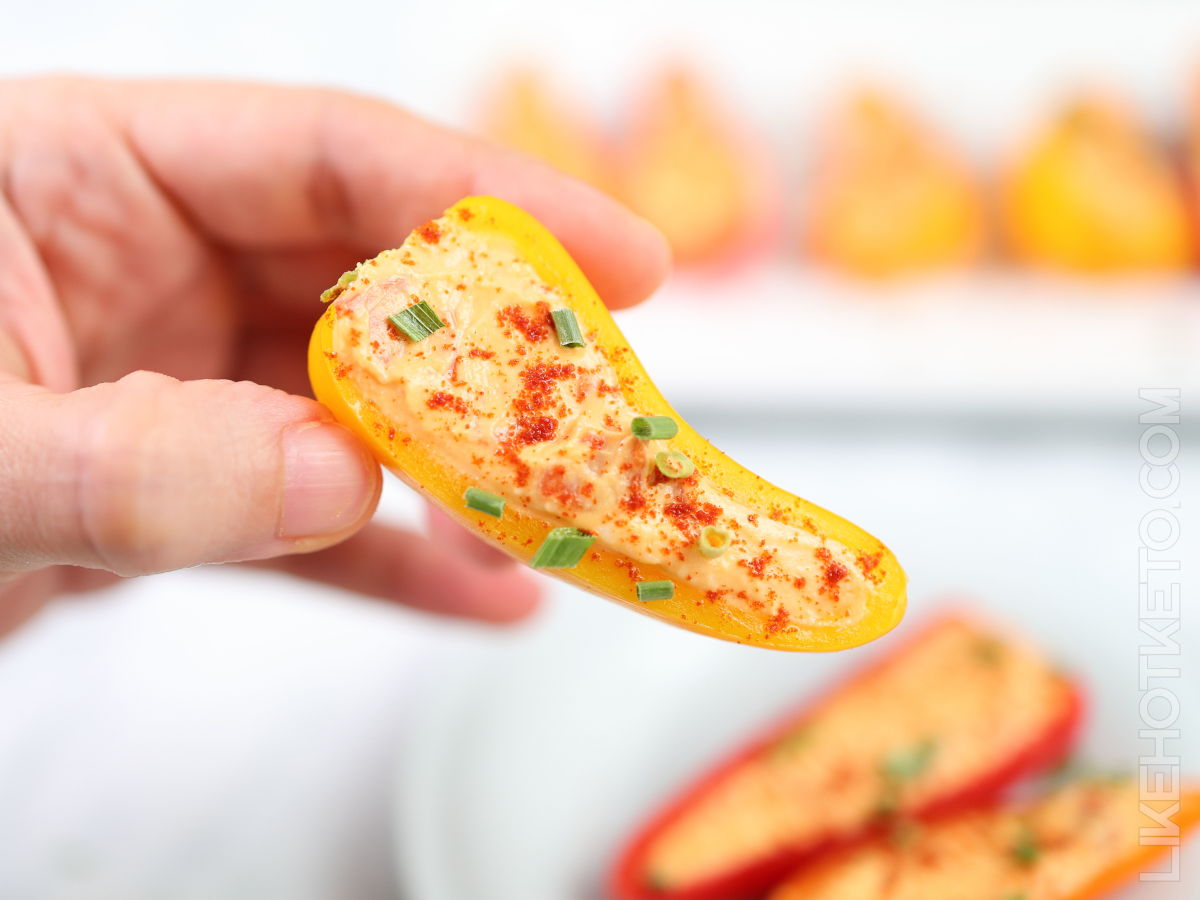 A hand holding a yellow mini bell pepper half filled with a creamy spread, topped with paprika and chopped chives.