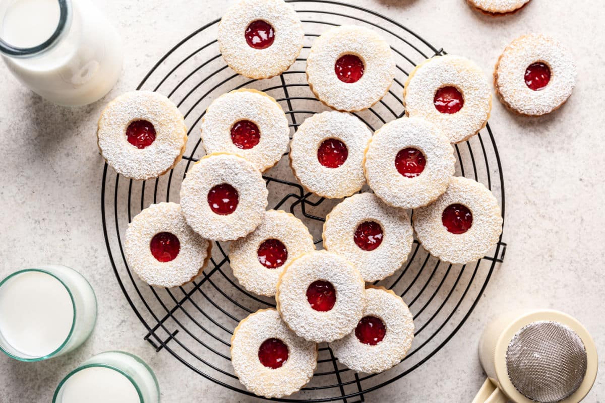 A cooling rack with powdered sugar-dusted sandwich cookies filled with red jam, surrounded by glasses of milk and a sifter on a light surface.
