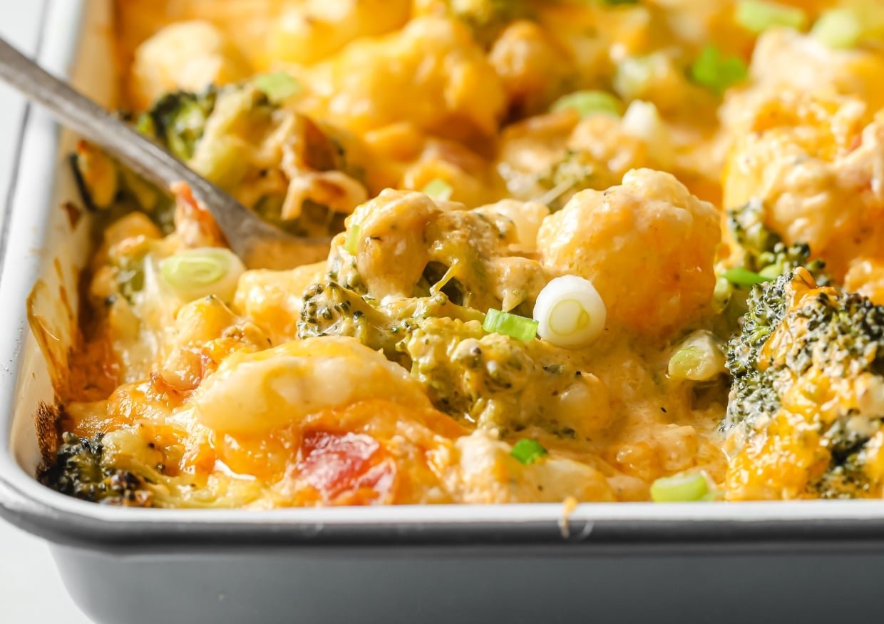 A close-up of a cheesy vegetable casserole with broccoli, cauliflower, and sliced green onions in a baking dish.