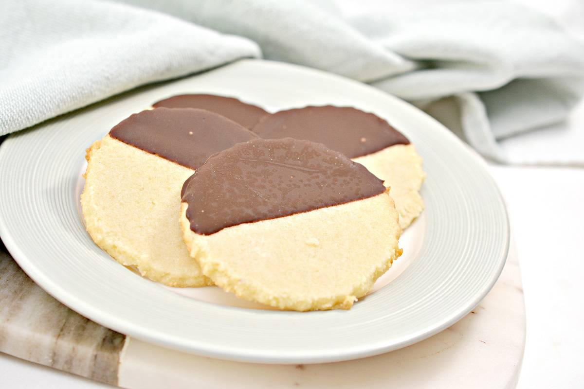Three round shortbread cookies partially dipped in chocolate are arranged on a white plate, with a light cloth in the background.