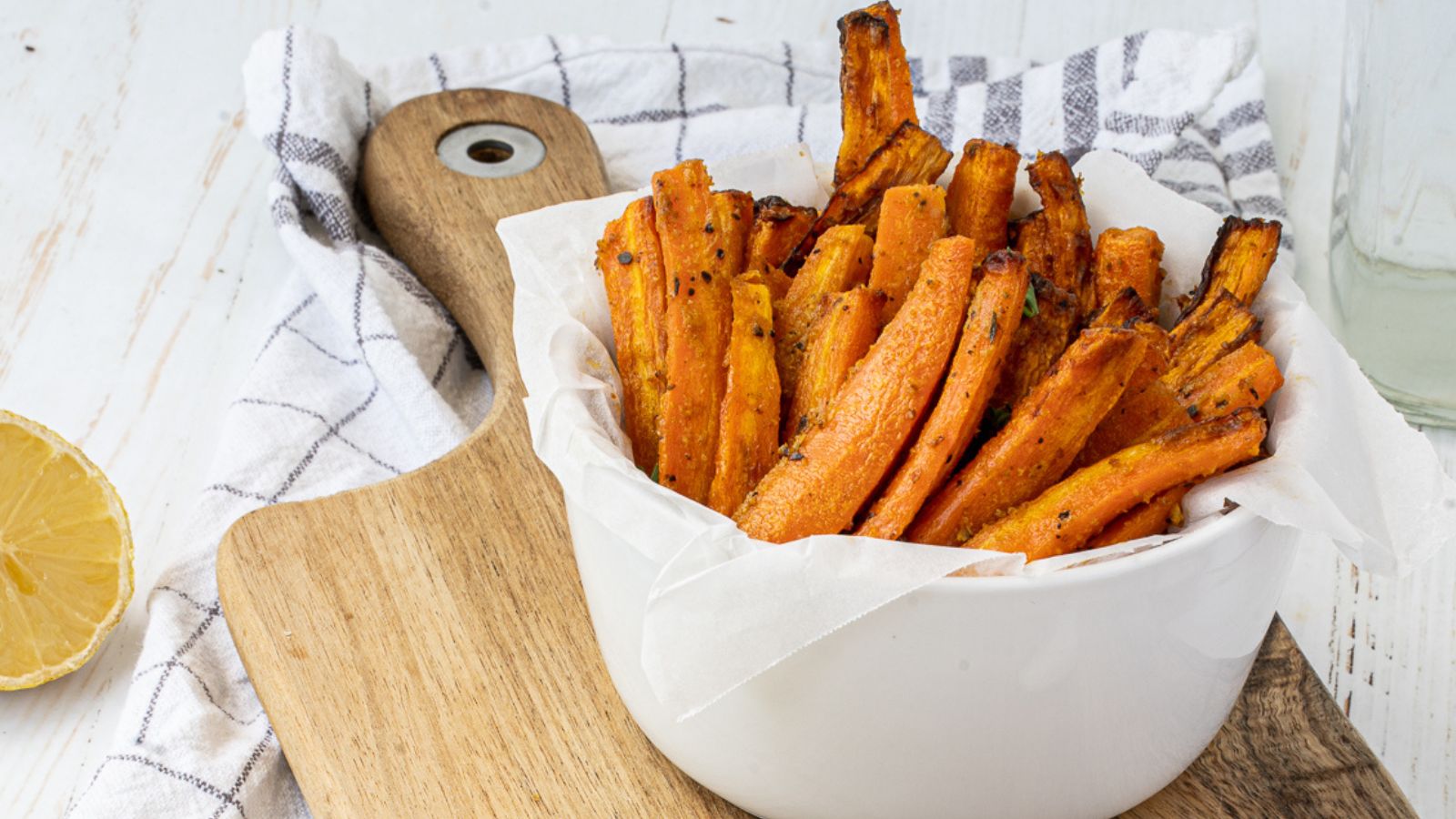 A white bowl filled with roasted carrot fries sits on a wooden board, with a checked cloth and a lemon half beside it.
