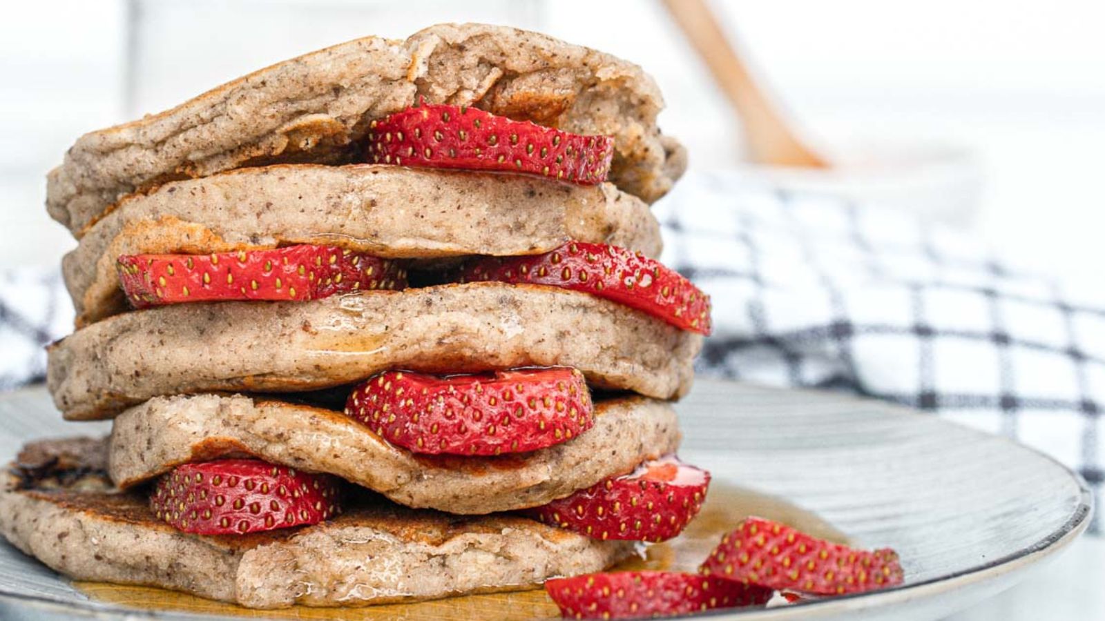 A stack of pancakes with sliced strawberries between each layer, served on a plate with a checkered cloth in the background.