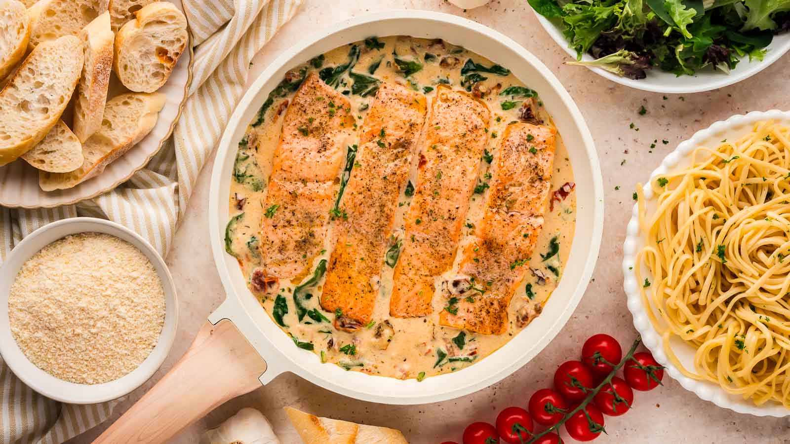 A skillet with creamy salmon fillets, spinach, and sauce, surrounded by bread slices, grated cheese, fresh salad, cherry tomatoes, and a bowl of cooked spaghetti.