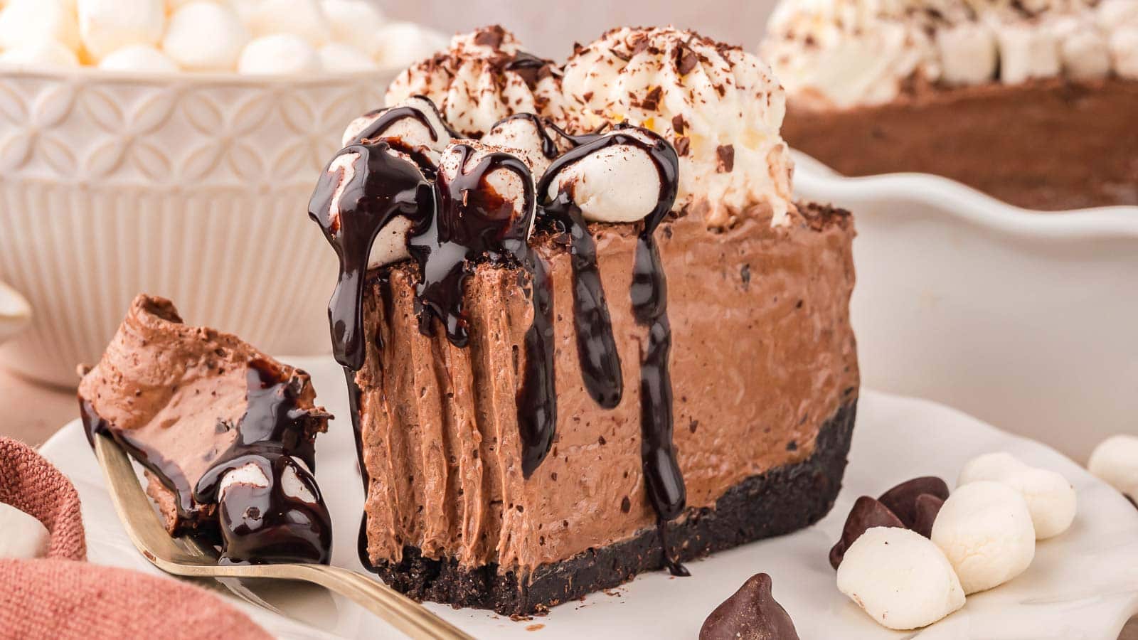 A slice of chocolate mousse pie with whipped cream, chocolate drizzle, and an Oreo cookie crust on a white plate with a fork.