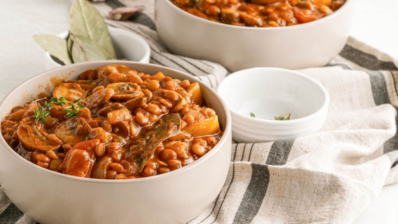 Two bowls of baked beans stew with vegetables and bay leaves sit on a striped cloth next to empty white bowls.