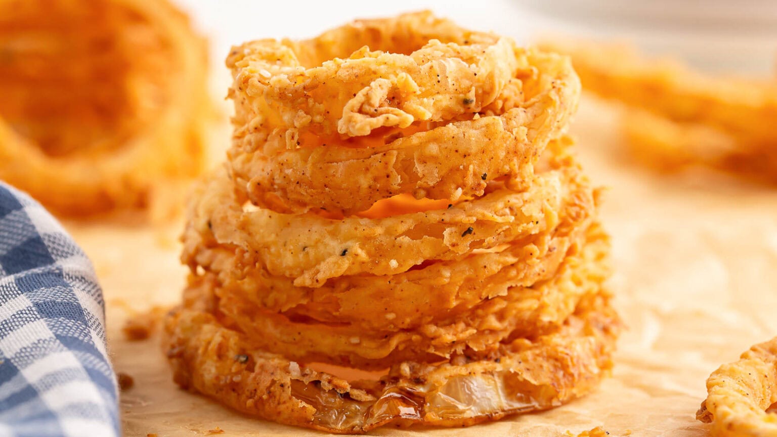 A stack of crispy, golden-brown fried onion rings sits on parchment paper next to a blue and white checkered cloth.