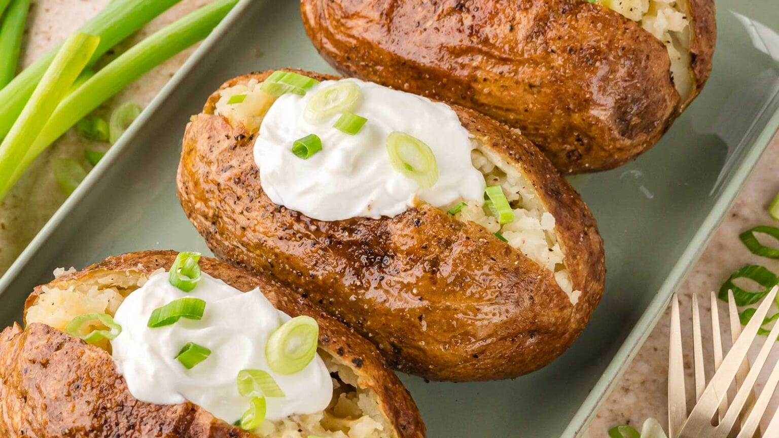 Three baked potatoes with crispy skins, split open and topped with sour cream and sliced green onions, served on a rectangular plate.