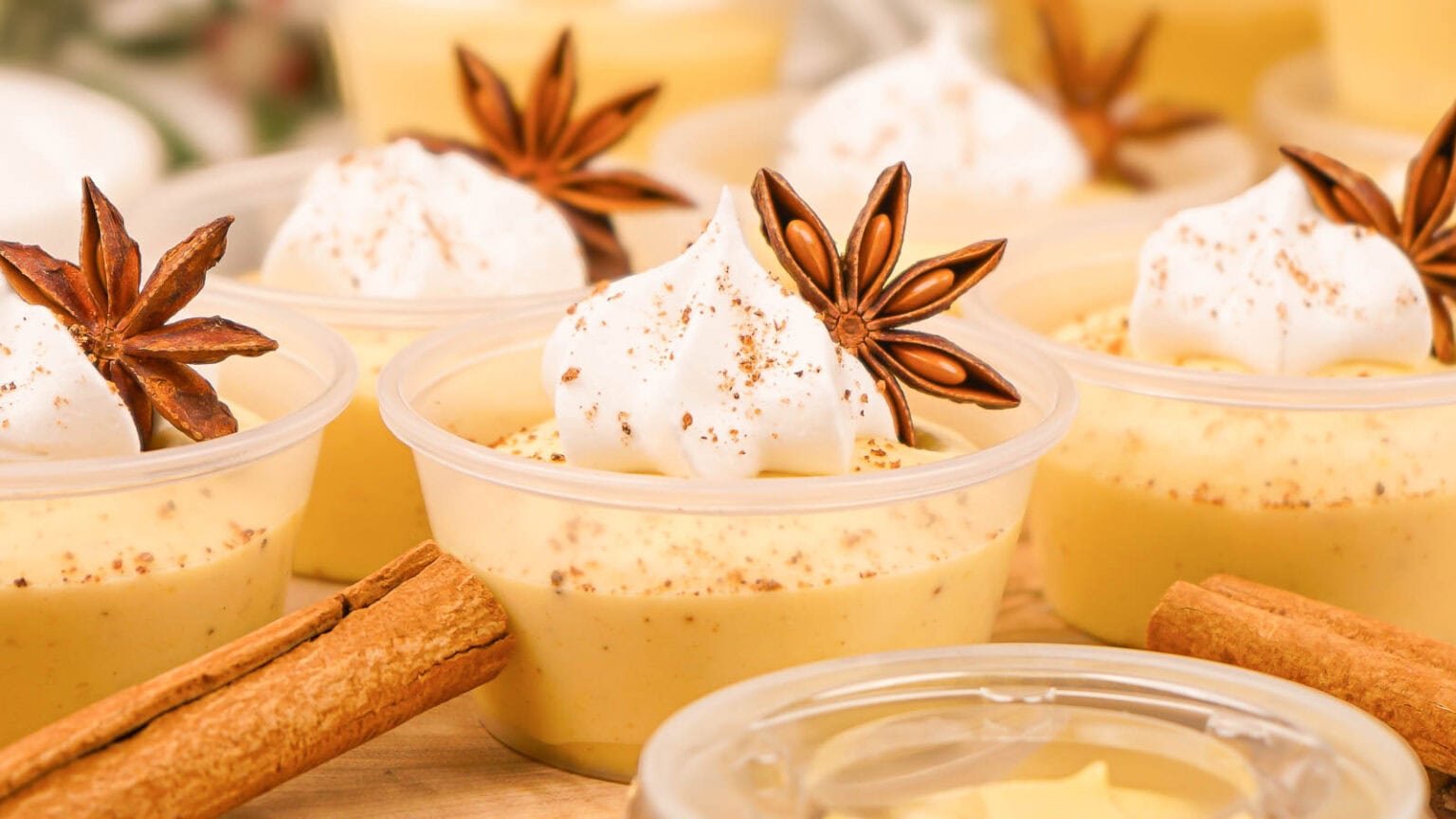 Small plastic cups filled with creamy yellow dessert, topped with whipped cream, star anise, and sprinkled spices, with cinnamon sticks nearby.