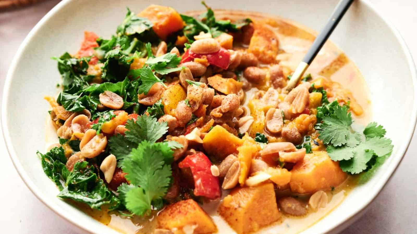 A bowl of vegetable curry with leafy greens, diced sweet potatoes, peanuts, cilantro, and a creamy sauce, served with a spoon.