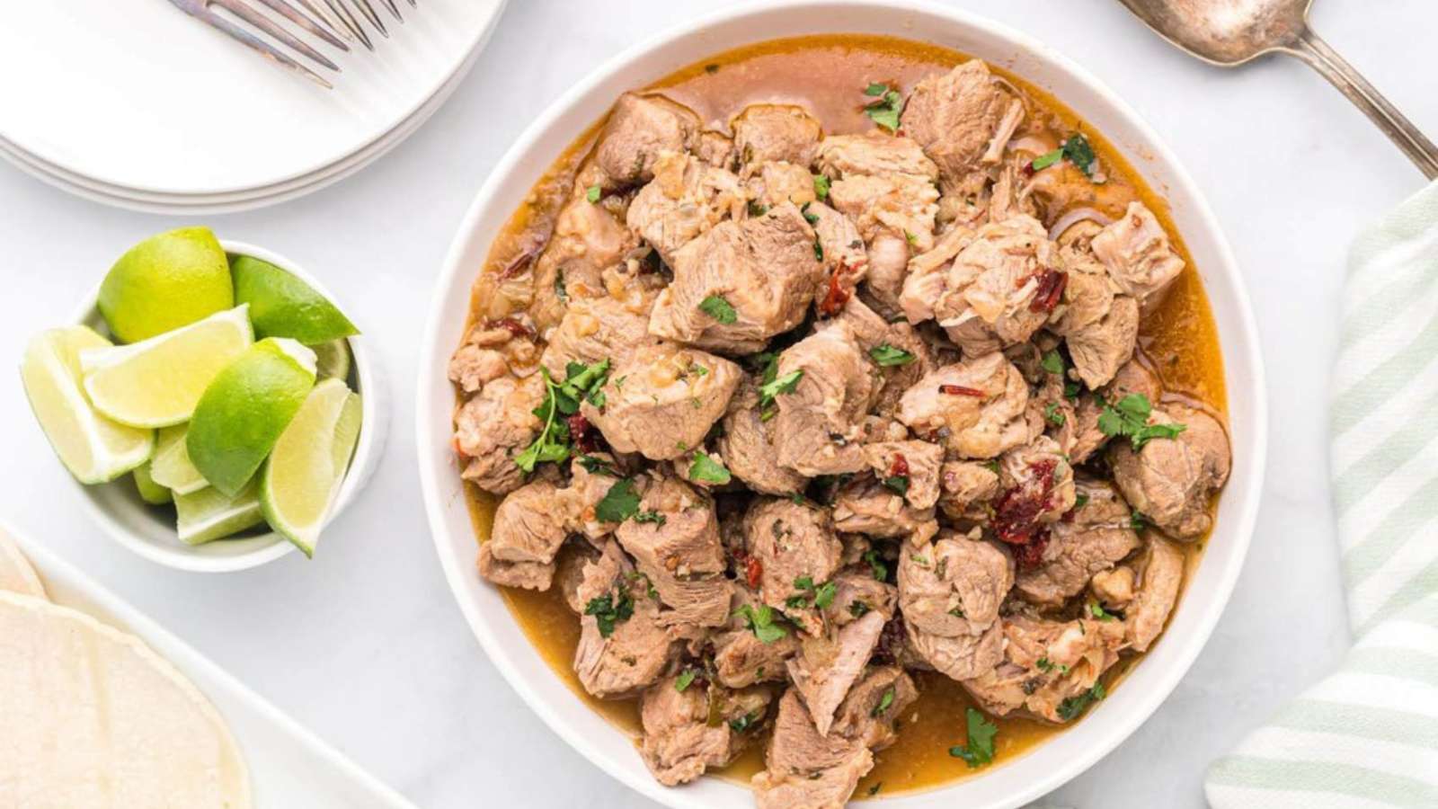 A bowl of cooked pork stew garnished with herbs, served with lime wedges on the side.