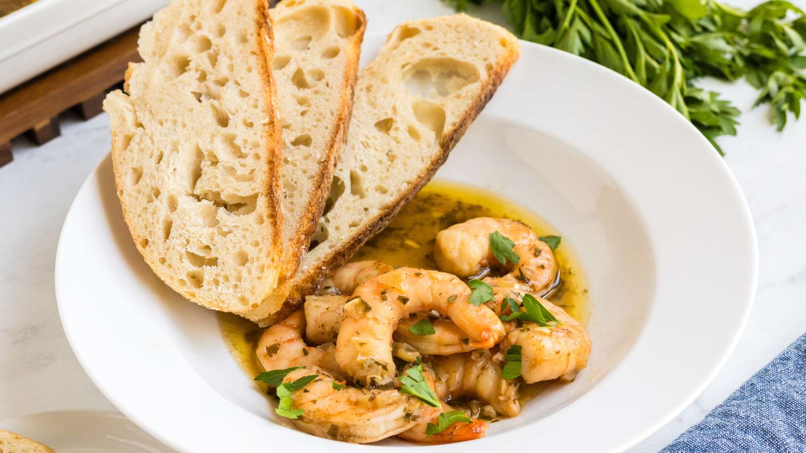 A white bowl containing shrimp in a seasoned sauce, garnished with herbs, served with two slices of rustic bread.