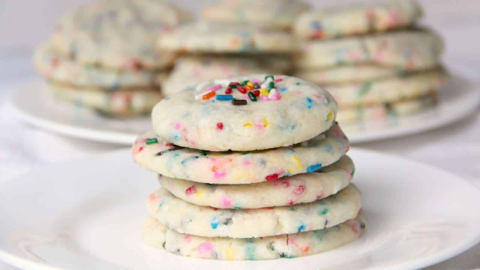 A stack of sugar cookies with colorful sprinkles sits on a white plate, with more cookies on plates in the background.