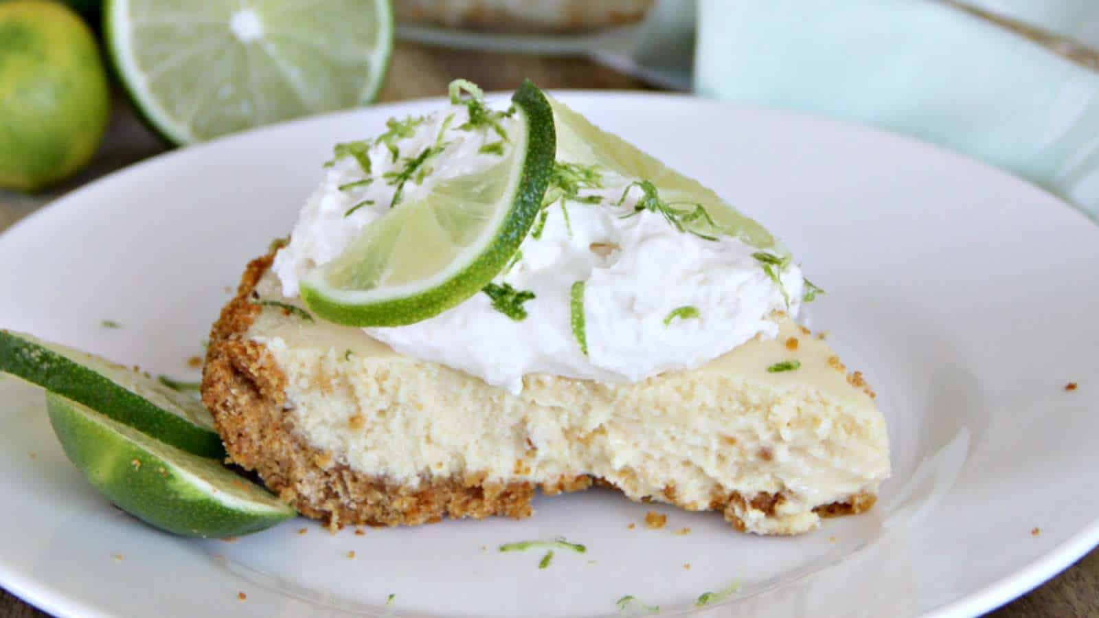 A slice of key lime pie topped with whipped cream and lime slices on a white plate, with a halved lime in the background.