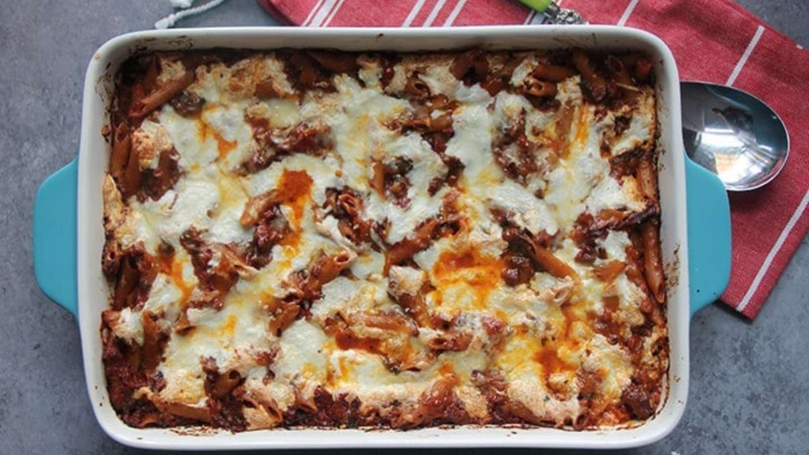 A baked pasta casserole topped with melted cheese in a rectangular dish, placed on a gray surface next to a red striped cloth and a metal spoon.