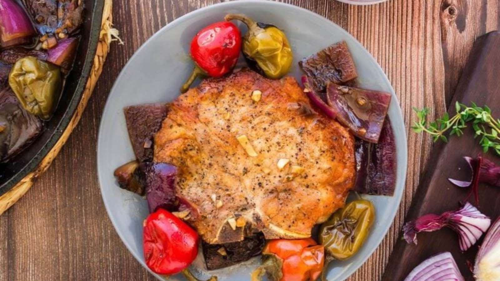 A cooked pork chop on a gray plate, surrounded by roasted red onions, red bell peppers, and green peppers on a wooden surface.