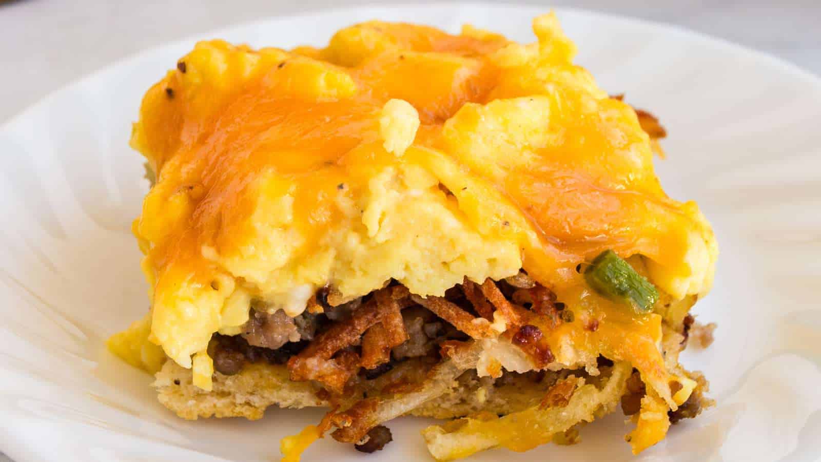 A slice of breakfast casserole with scrambled eggs, sausage, cheese, and hash browns on a white plate.