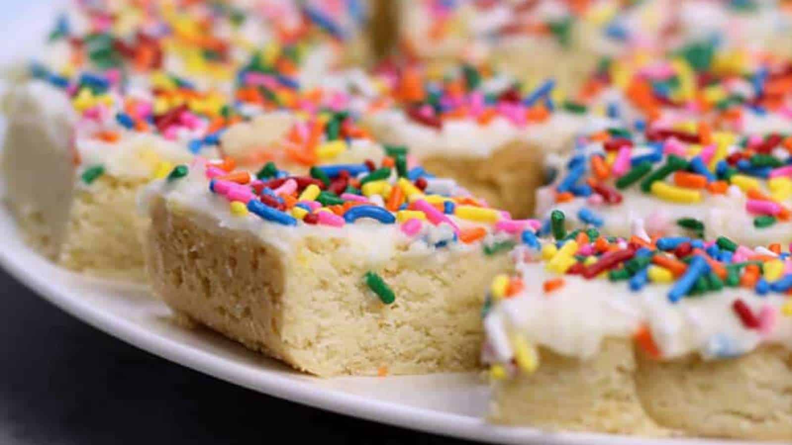 Frosted sugar cookie bars topped with colorful rainbow sprinkles arranged on a white plate.