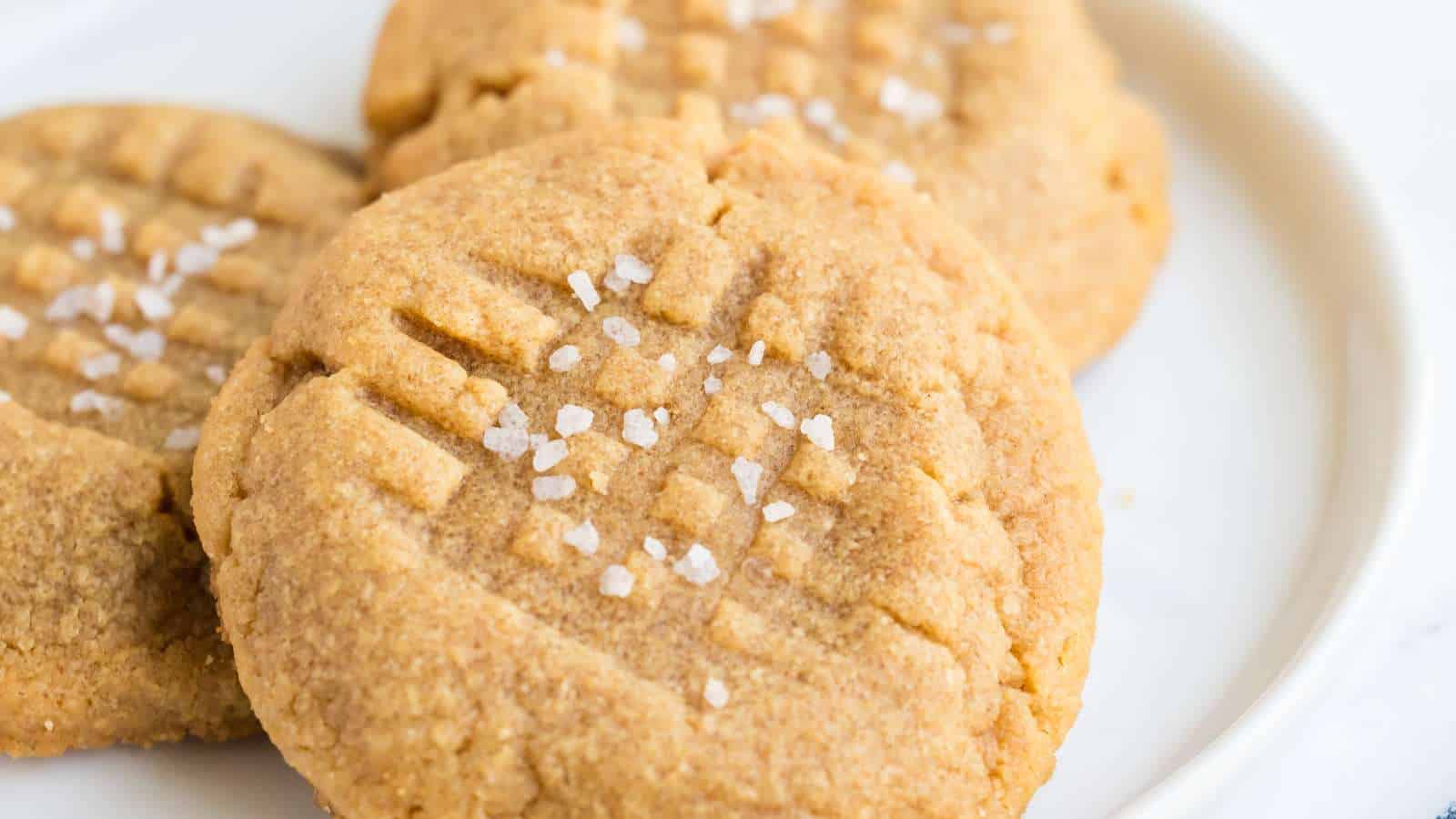 Three peanut butter cookies with a crisscross fork pattern and coarse salt on top, arranged on a white plate.