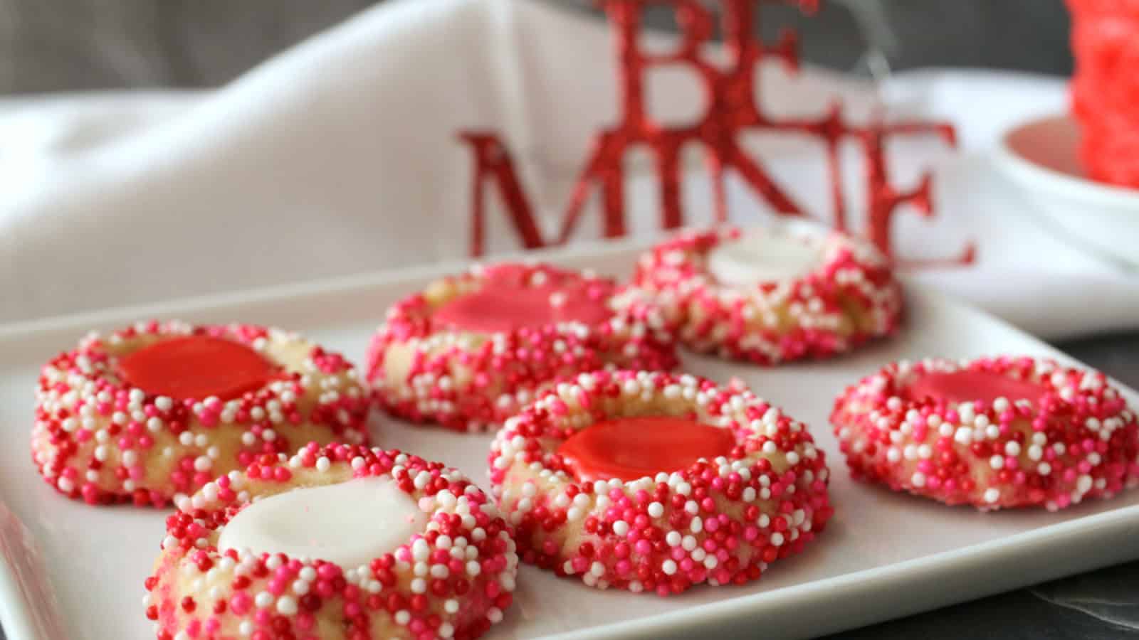 A plate of round cookies topped with red and white icing and coated in red, pink, and white sprinkles. A Be Mine decoration is in the background.