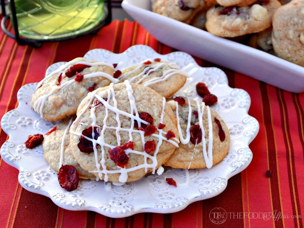 A plate of cookies with white icing drizzle and dried cranberries sits on a red striped tablecloth, with more cookies in a white dish in the background.