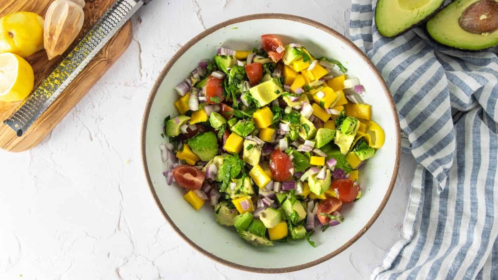 A bowl of chopped salad with avocado, mango, tomatoes, red onion, and cilantro, placed on a white surface next to half an avocado, lemon, and a striped cloth.