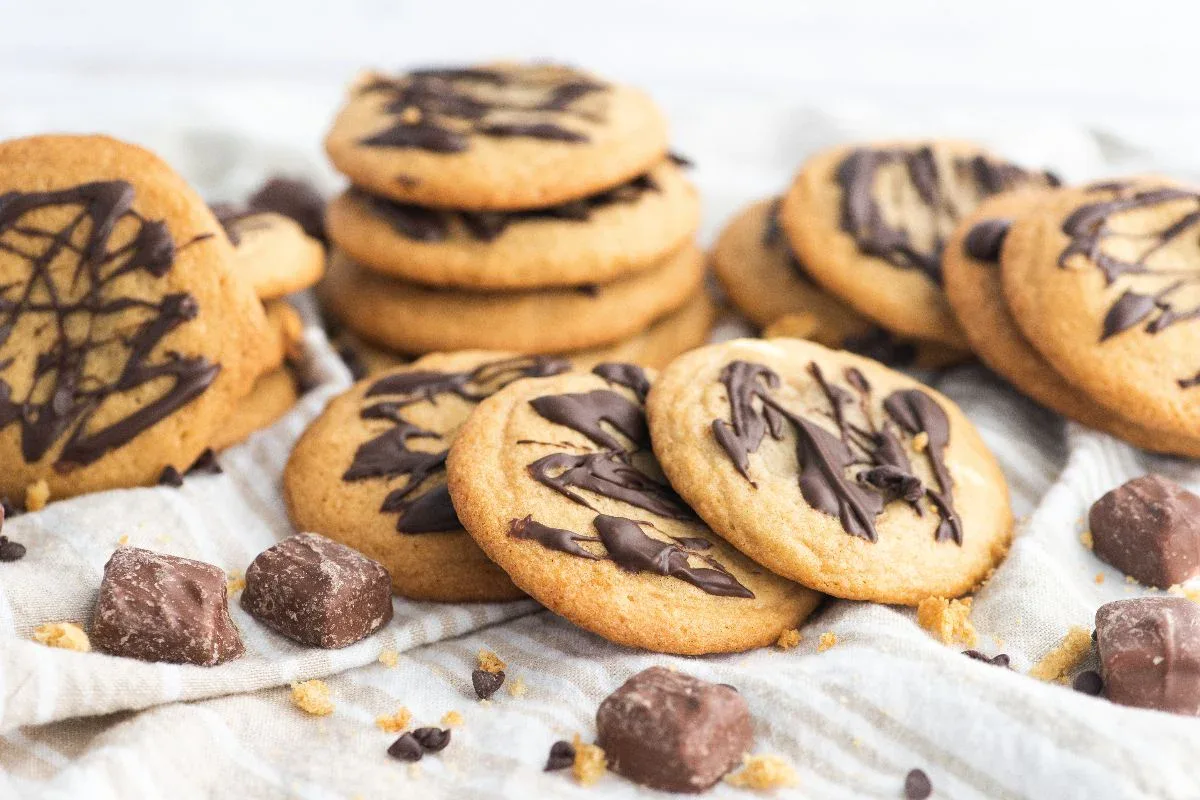 A group of chocolate-drizzled cookies arranged on a cloth with small chocolate pieces scattered around.