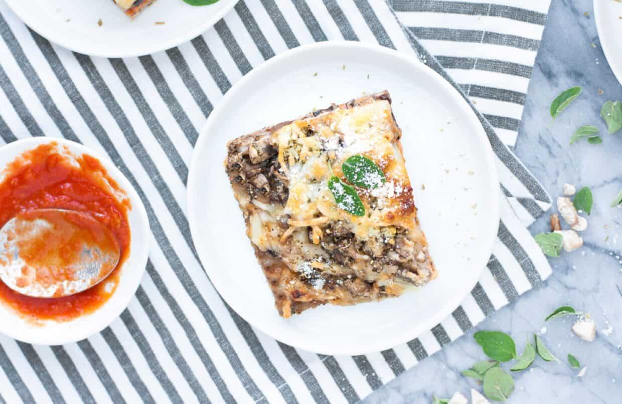 Overhead view of a slice of lasagna topped with cheese and herbs on a white plate, next to a bowl of tomato sauce and scattered fresh oregano leaves.