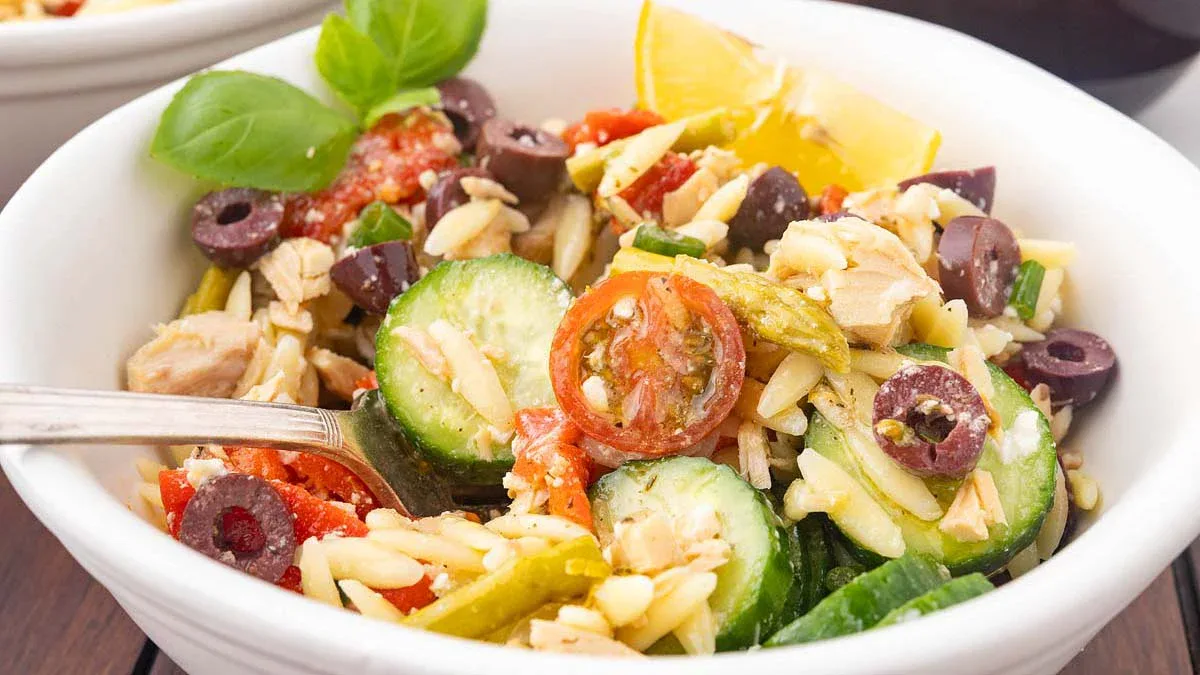 A bowl of orzo pasta salad with cherry tomatoes, sliced cucumber, olives, basil, tuna, and a lemon wedge.