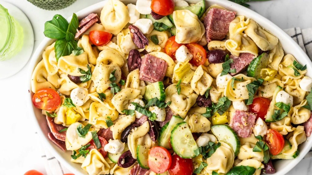 A bowl of tortellini pasta salad with cherry tomatoes, sliced cucumbers, mozzarella balls, olives, pepperoni, and fresh basil.
