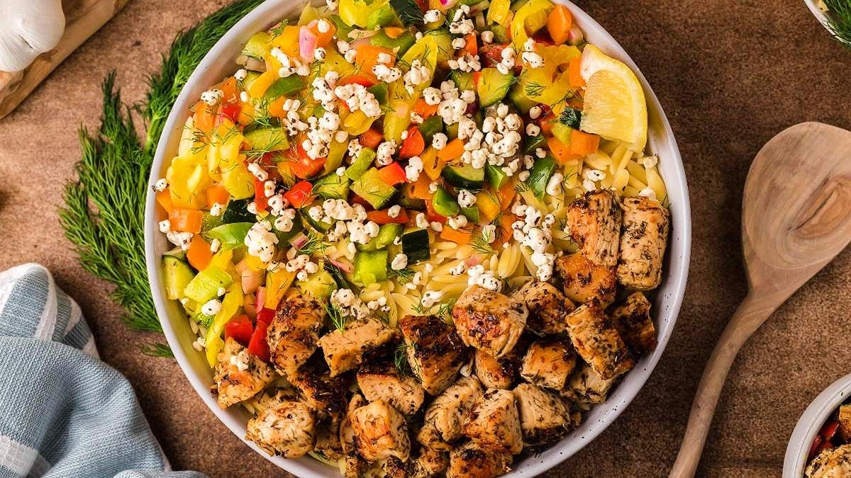 A bowl filled with orzo pasta, diced colorful vegetables, grilled chicken pieces, crumbled cheese, and a lemon wedge, arranged on a brown surface with herbs and a wooden spoon nearby.